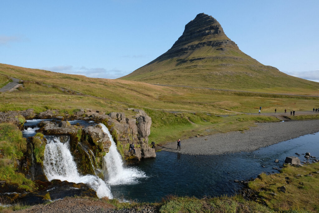 Im Vordergrund des Bildes sind die Kirkjufellsfoss-Wasserfälle mit klar sprudelndem Wasser zu sehen, während im Hintergrund der markante Berg Kirkjufell aufragt. Das Bild zeigt eine weite natürliche Landschaft mit grünen Wiesen und einem ruhigen, unberührten Ambiente.