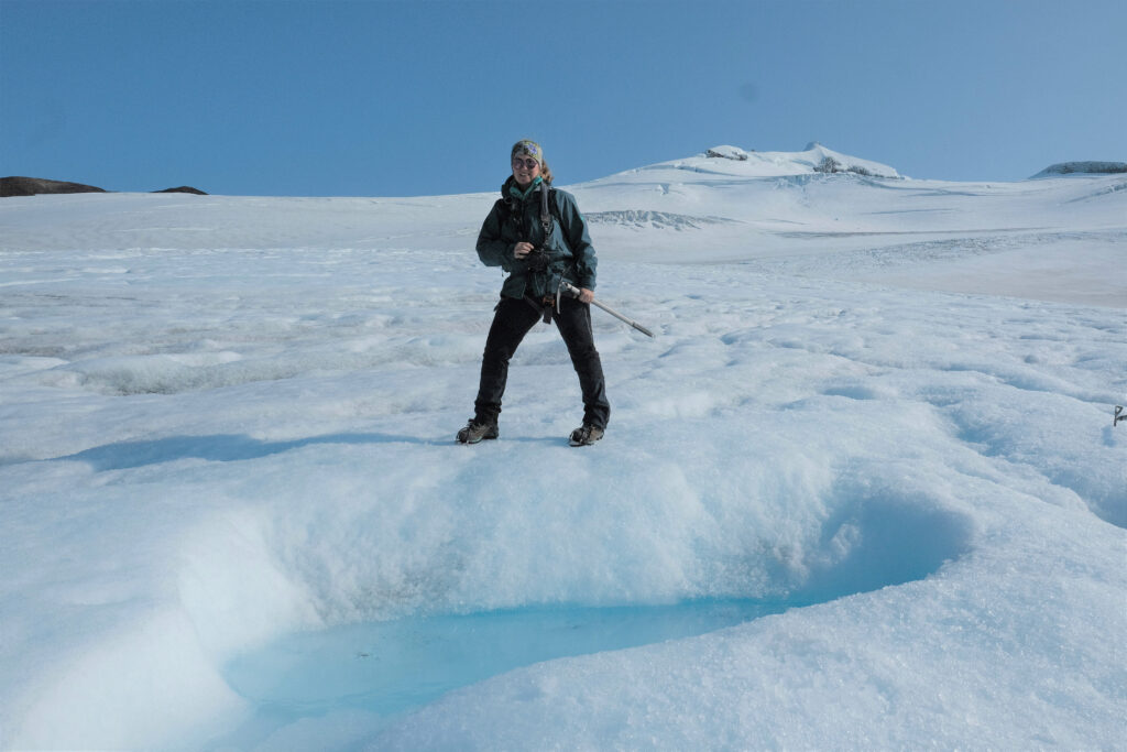Weibliche Person mit Wanderkleidung und Eispickel steht auf einem verschneiten Gletscher mit einer kleinen blauen Schmelzwasser-Pfütze, umgeben von einer weiten, schneebedeckten Landschaft und blauem Himmel.